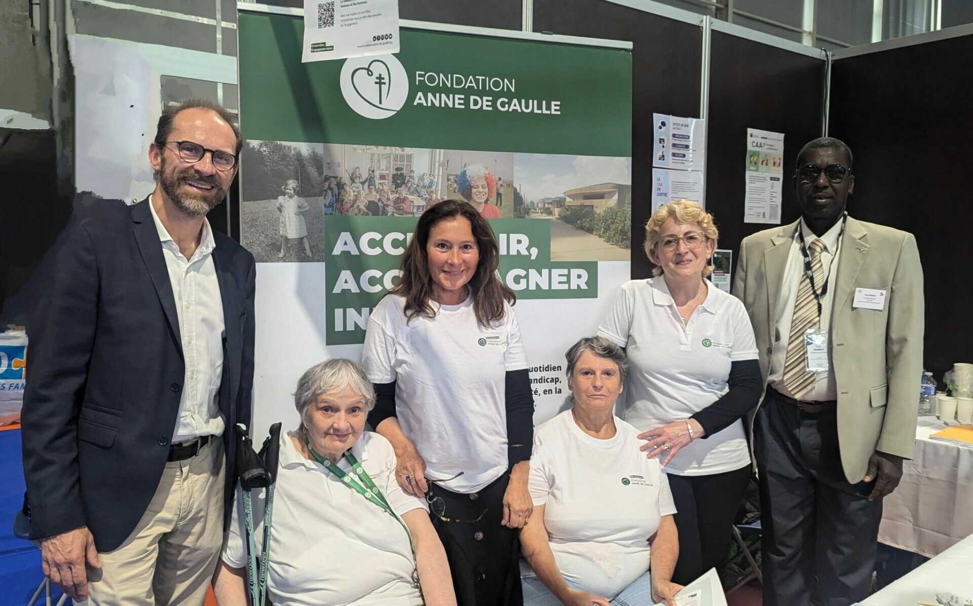 L’image montre six personnes debout devant une bannière de la “FONDATION ANNE DE GAULLE”. La bannière est blanche avec des lettres bleues et comporte les mots “ACCUEILLIR, ACCOMPAGNER” en dessous du nom de la fondation. Les personnes présentes semblent être des participants ou des représentants de l’événement, et leurs visages sont masqués par des rectangles marron pour préserver leur vie privée. L’événement semble se dérouler dans un espace intérieur, probablement une salle d’exposition ou un hall, comme le suggèrent les structures en arrière-plan qui ressemblent à des stands ou des kiosques. L’éclairage est lumineux, indiquant qu’il s’agit d’un environnement bien éclairé, typique des événements publics ou des expositions. Les personnes sur la photo sont habillées de manière décontractée mais soignée, ce qui est courant pour ce type d’événement. Il est possible que l’événement soit lié à des activités de soutien ou d’accompagnement, en accord avec la mission de la Fondation Anne de Gaulle, qui se concentre sur l’accueil et l’accompagnement des personnes dans le besoin.